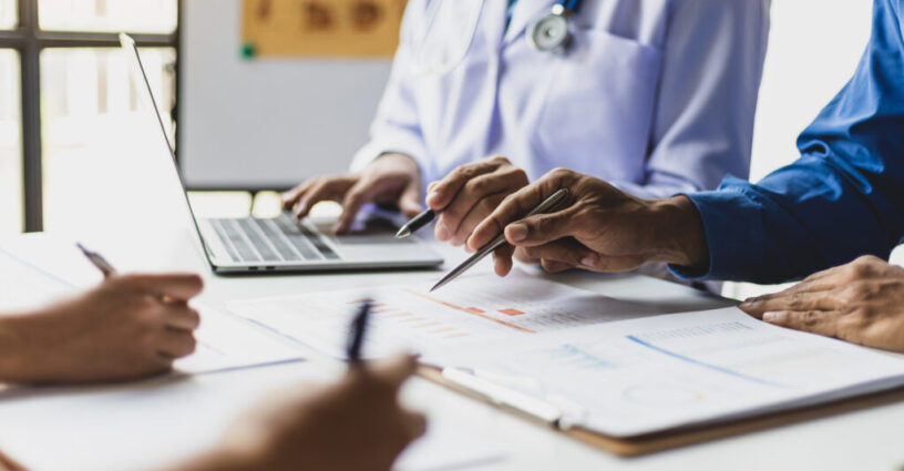 Team, medical analysts and doctors consulting with paperwork of graphs, data and charts in hospital conference room. Healthcare staff discussing statistics, results of research and innovation.